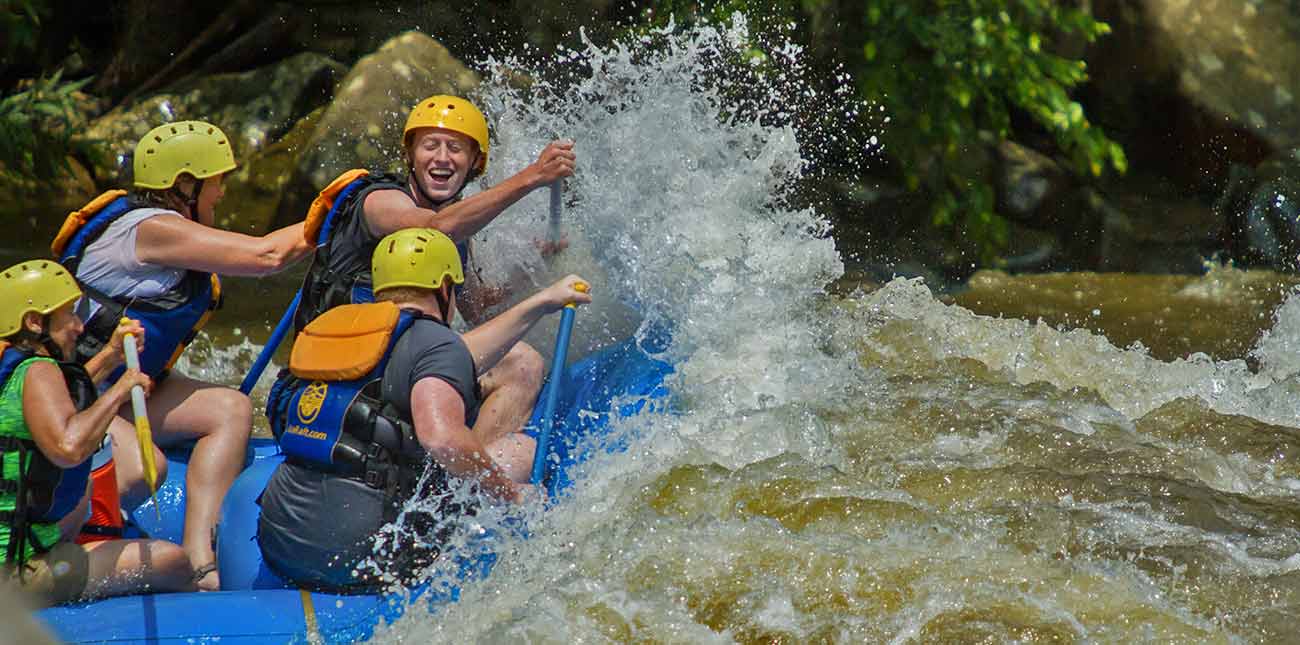 A huge wave crashes over the bow of a raft while it's riders smile.